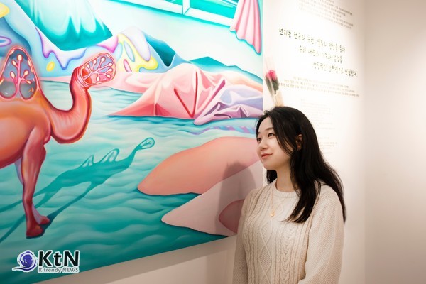 넌지 작가가 전시장에서 자신의 대형 작품 앞에 서 있다. 전구와 신경세포를 연상시키는 독특한 형상의 낙타와 파스텔톤의 몽환적인 풍경이 어우러진 작품은 현실과 상상의 경계를 넘나드는 작가만의 예술적 상상력을 보여준다. 사진 제공=넌지 작가 / K trendy NEWS DB ⓒ케이 트렌디뉴스 무단전재 및 수집, 재배포금지